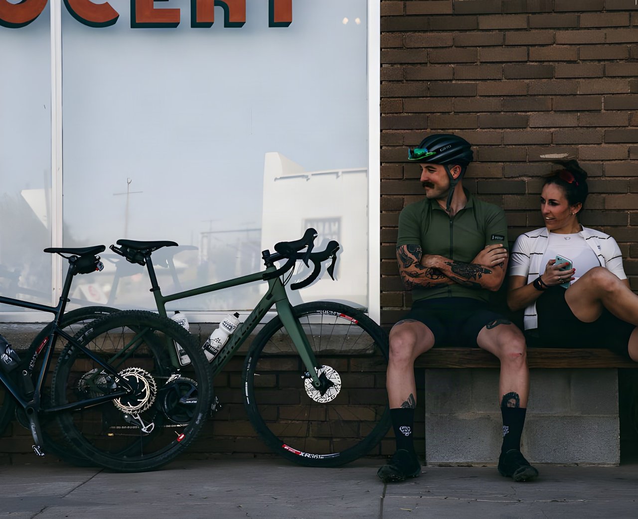 Two riders rest on a bench outside a shop beside a green Santa Cruz Stigmata and a second gravel bike.