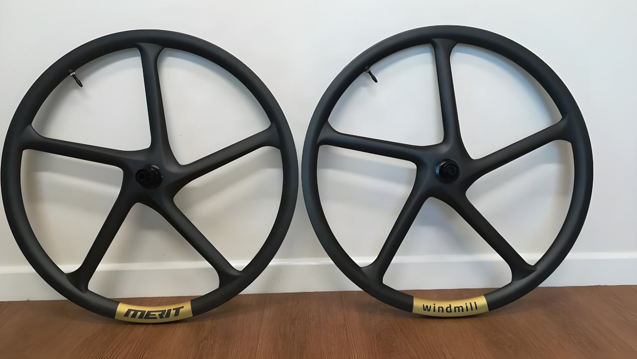 Pair of Merit Windmill five-spoke carbon gravel wheels with gold rim decals, leaning against a wall.