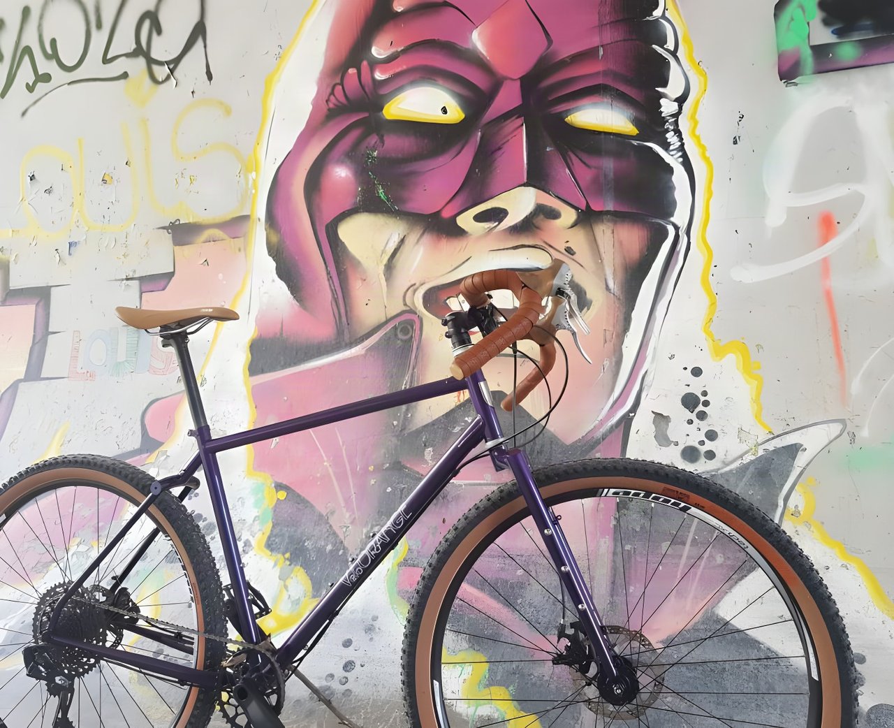 Purple Velo Orange Piolet with tan-wall tyres and brown bar tape leaning against a graffiti mural.