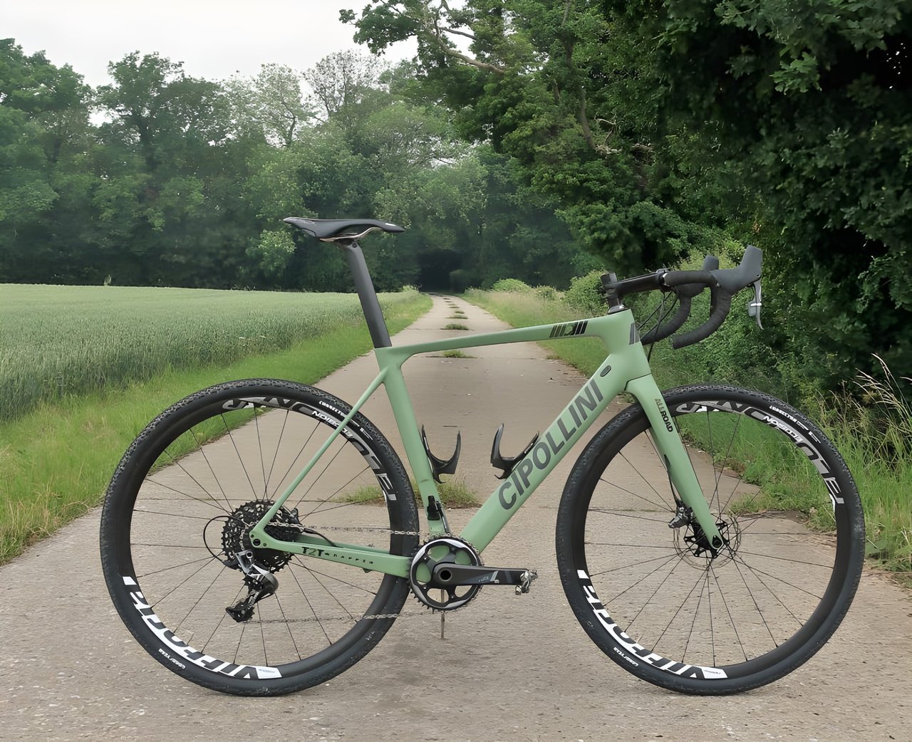 Cipollini MCM All Road gravel bike in matte army green, side view on a concrete farm track beside a green crop field.