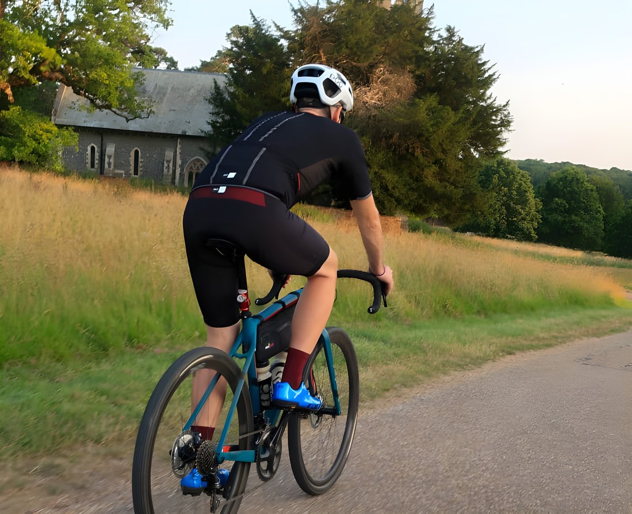 Rider in PEdALED Odyssey jersey and bibshorts on a teal gravel bike, passing a stone church on a quiet lane.