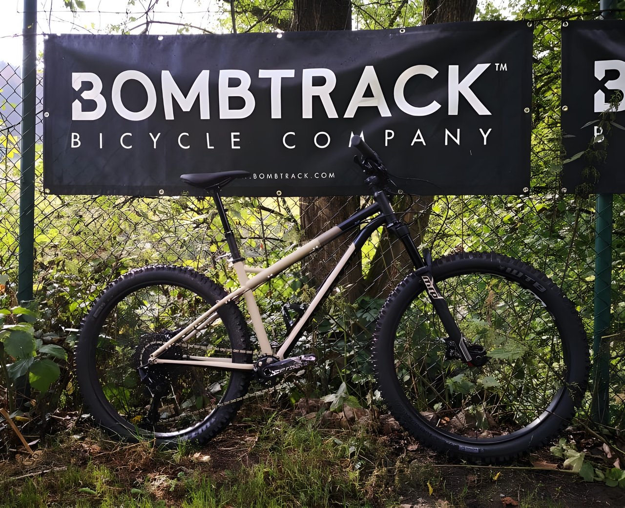 Bombtrack Cale 27.5+ hardtail adventure bike in sand and black, leaning on a fence below the brand's banner.