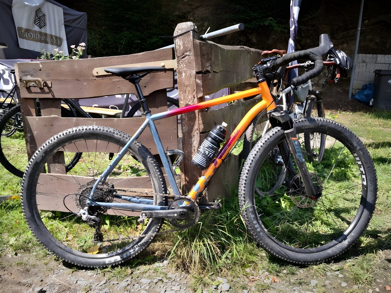 Orange-to-blue fade Bombtrack Hook ADV 2020 with air-sprung fork and MTB tyres, propped against a wooden fence.