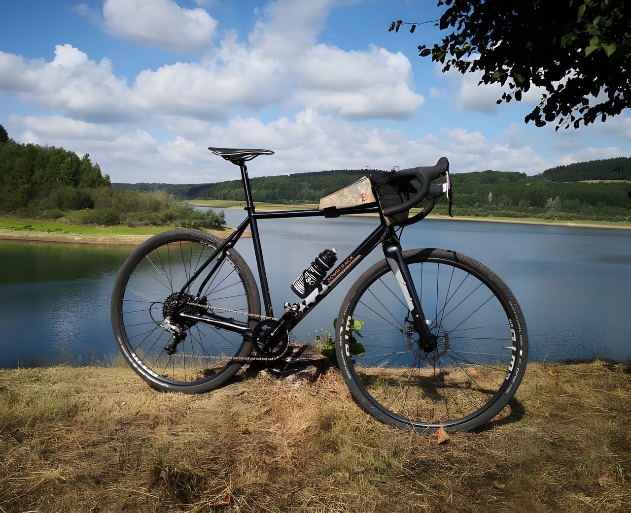 Black Bombtrack Hook 2 gravel bike with a top-tube bag, parked on the shore of a reservoir under a blue sky.