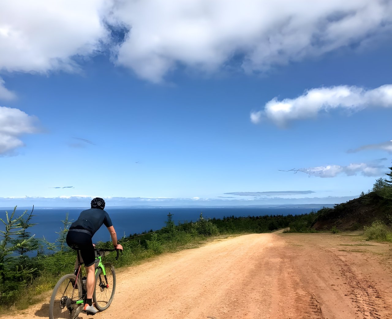 Rider on a green gravel bike climbs a sandy track above a deep blue sea on a summer gravel holiday.