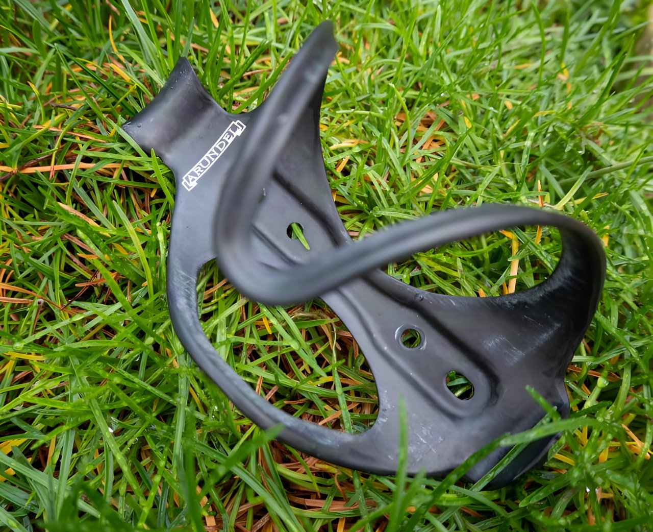 Arundel DTR/STR carbon bottle cage lying on grass, showing its side-entry shape and mounting holes.