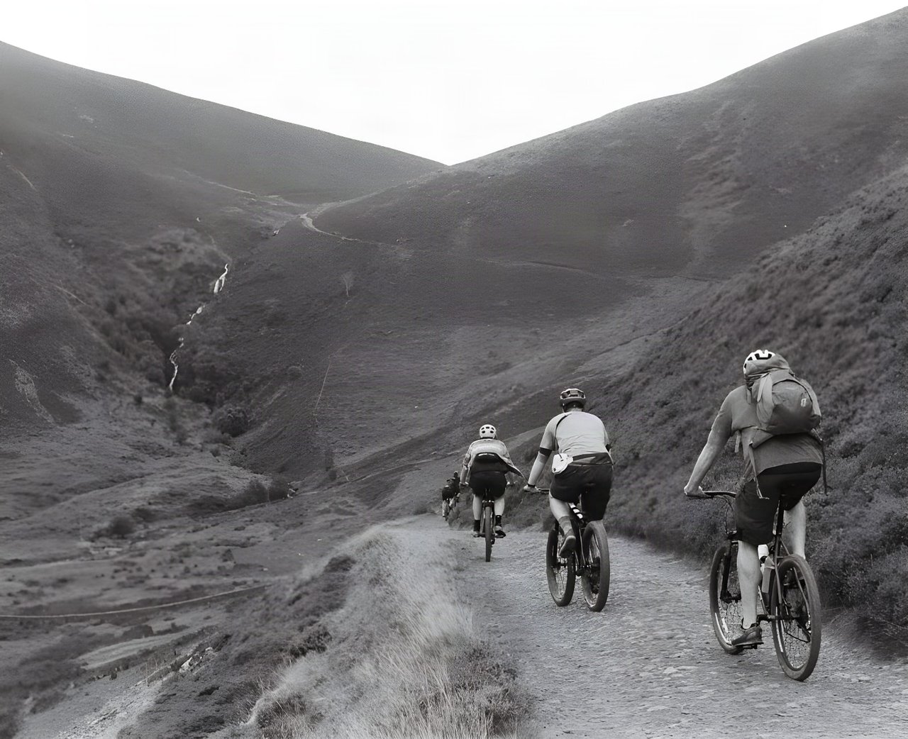 Three riders climb a gravel track up a steep-sided Lake District valley on the Low Key Lakes Weekender, in black and white.