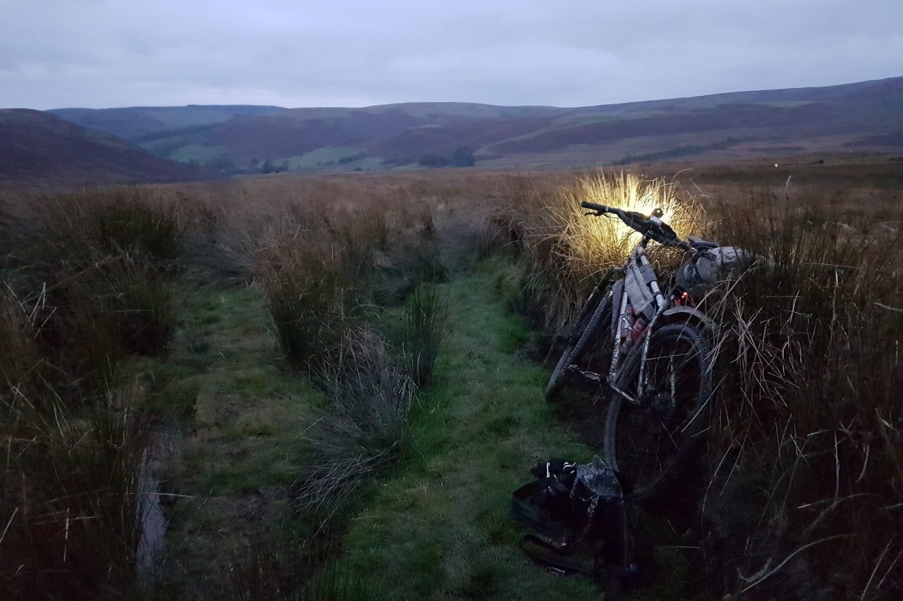 Loaded bikepacking bike with its front light on, propped in tall grass on a Welsh moorland track at dusk, Bear Bones 200.