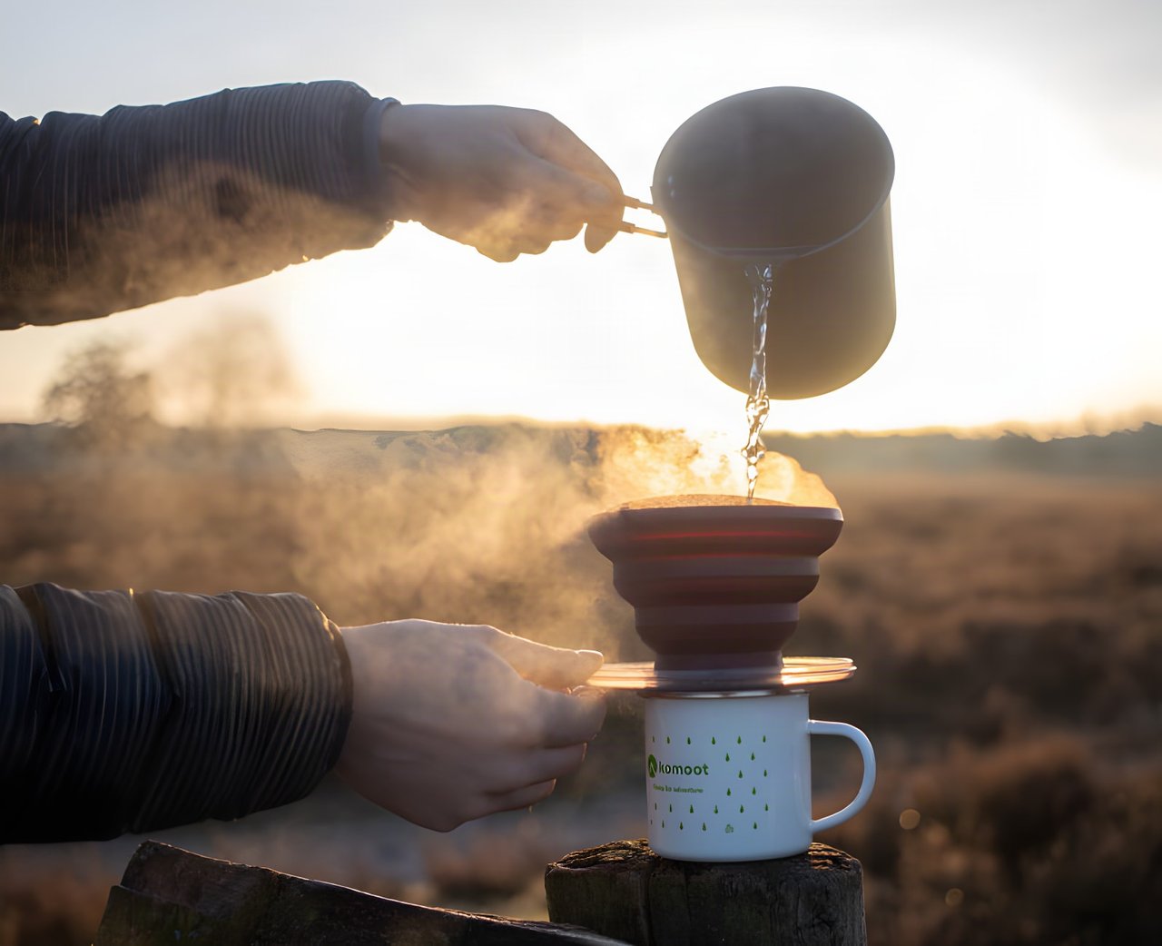 Hot water poured from a pot into a pour-over filter on a komoot mug at sunrise on a Dutch daypacking coffee stop.