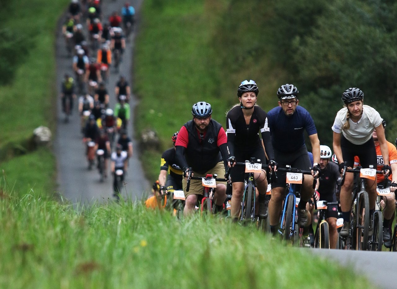 Large pack of Dirty Reiver riders with handlebar number boards climbs a narrow lane through green hills in Kielder Forest.