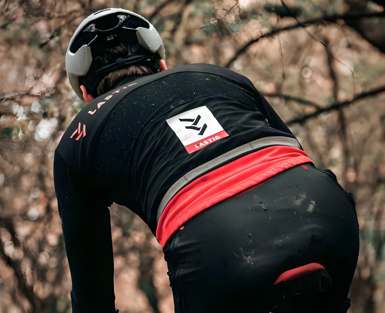 Rear view of a rider in a black LASTIG cyclocross jacket and tights, splashed with mud, in autumn woodland.