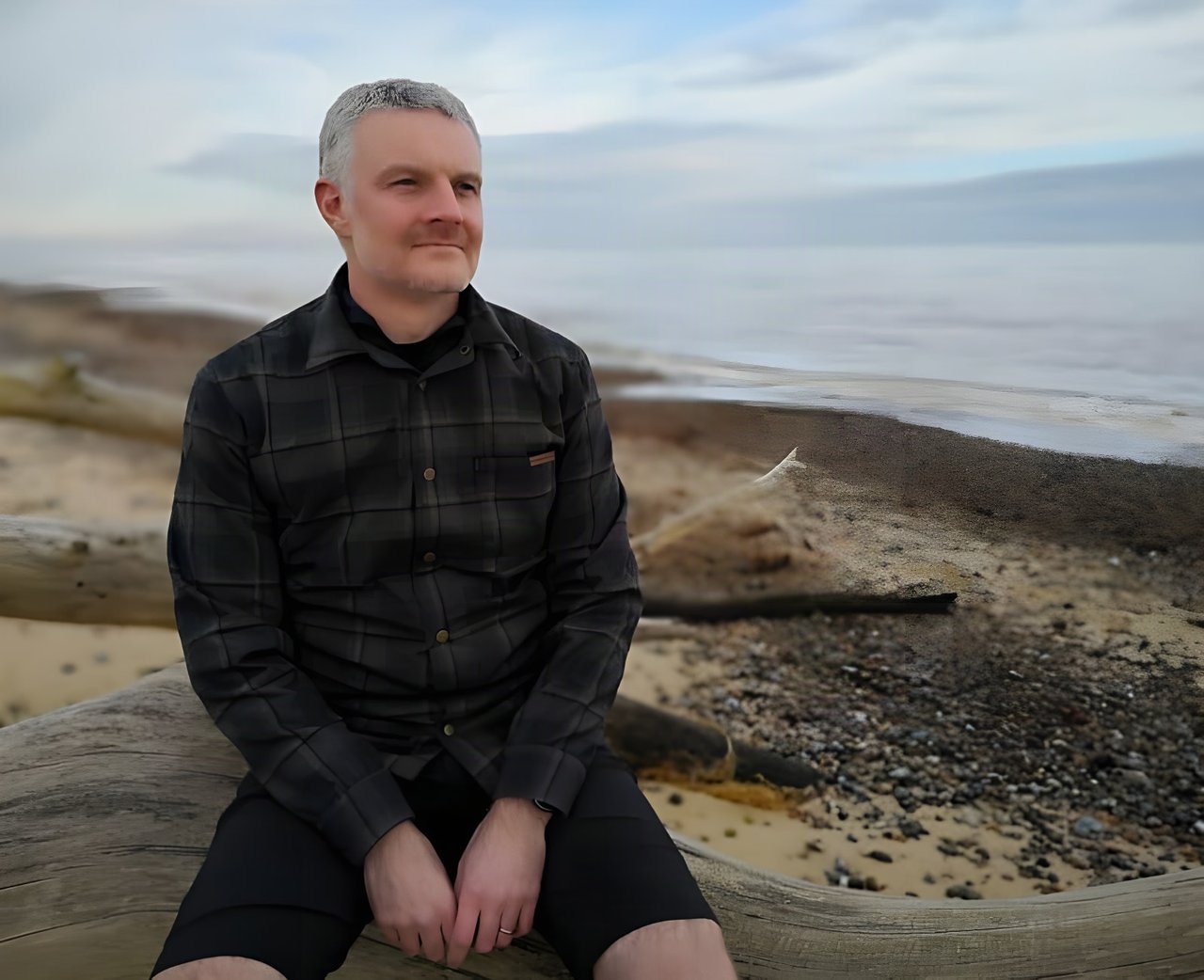 Rider in the dark plaid Endura Hummvee Shacket sits on driftwood on a pebbly beach, looking out to sea.