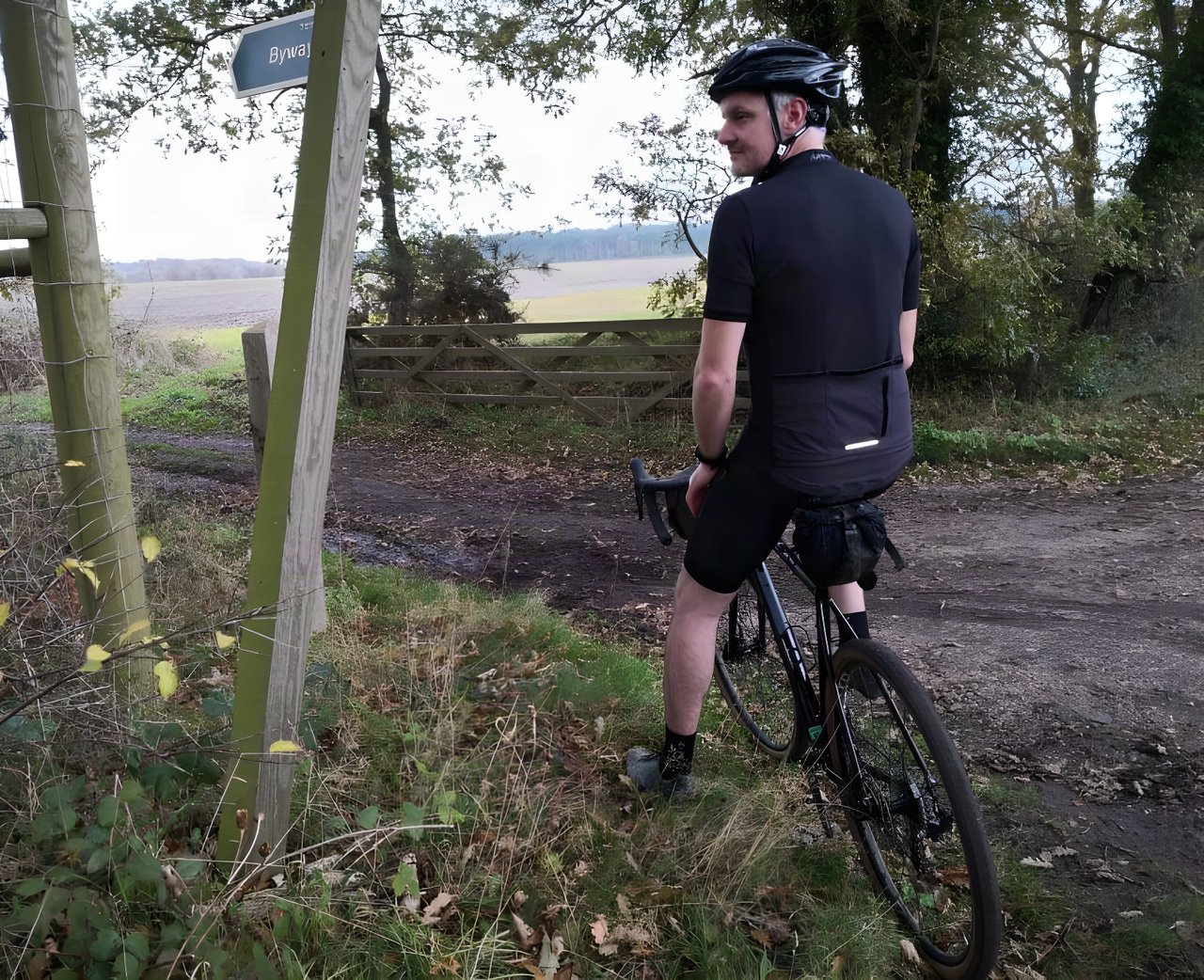 Rider in the black Endura MTR Adventure Jersey pauses on a gravel bike beside a byway sign and a farm gate.