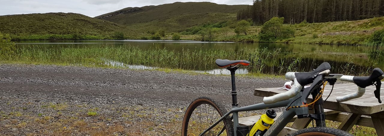 Grey gravel bike at a picnic table by a reedy lake and wooded hills in Fermanagh, home of the Lakelander Gravel Grinder.