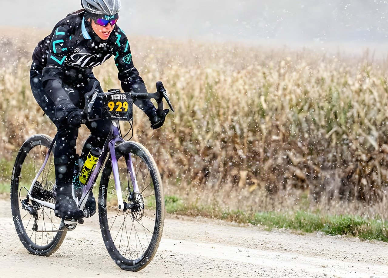 Rider on a purple-fade All-City Cosmic Stallion racing along a gravel road in falling snow beside a cornfield.