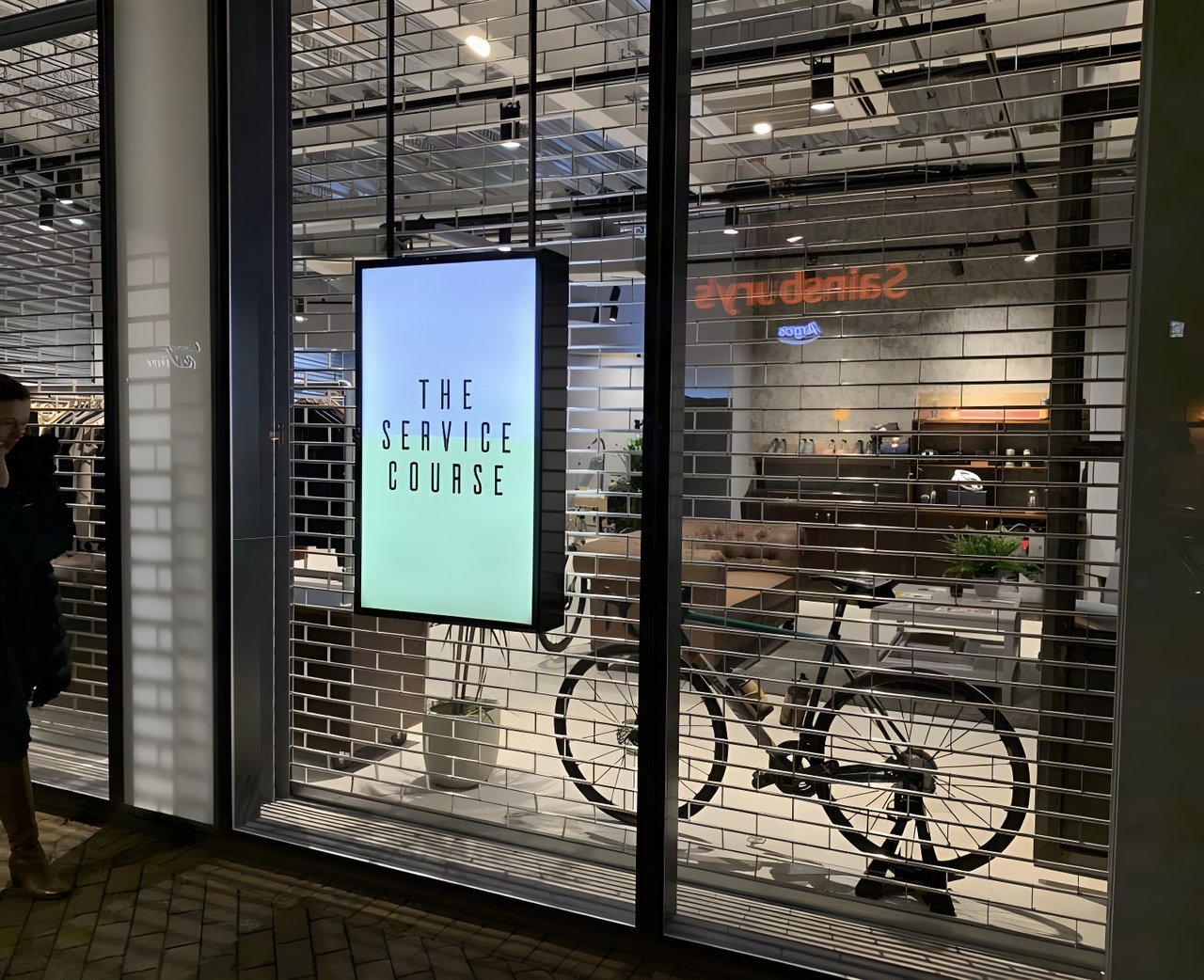 The Service Course Wilmslow shopfront after dark, with an illuminated sign and a bike visible behind the shutter.