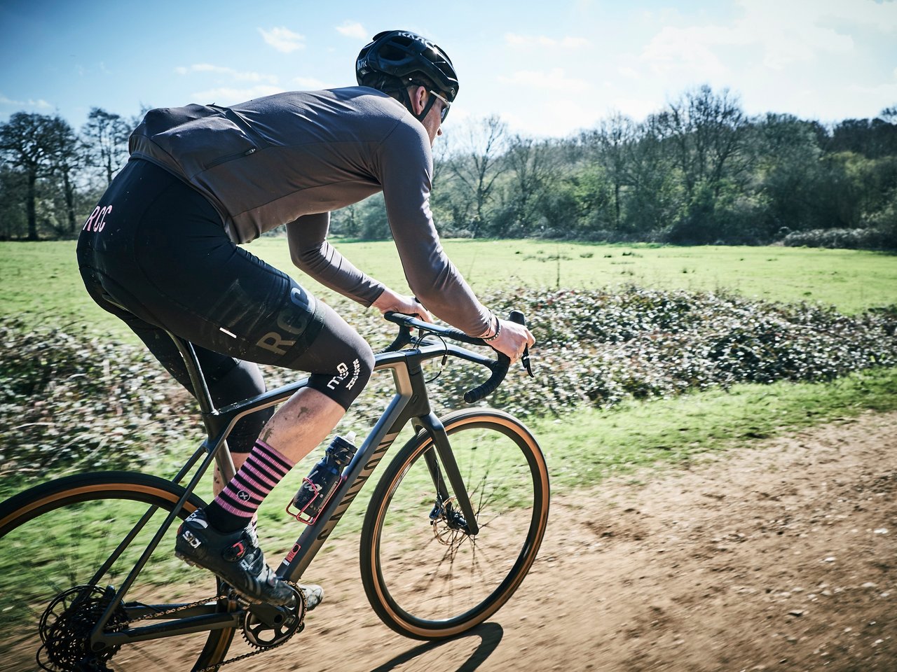 North London Dirt 3 rider on a Canyon gravel bike speeds along a dry dirt track between fields in bright sunshine.