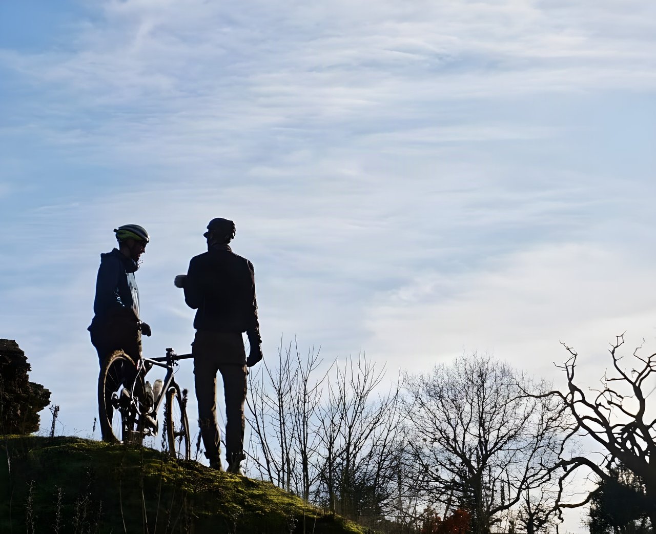 Two cyclists in silhouette chat beside a bike on a grassy rise, for the Unpaved Podcast.