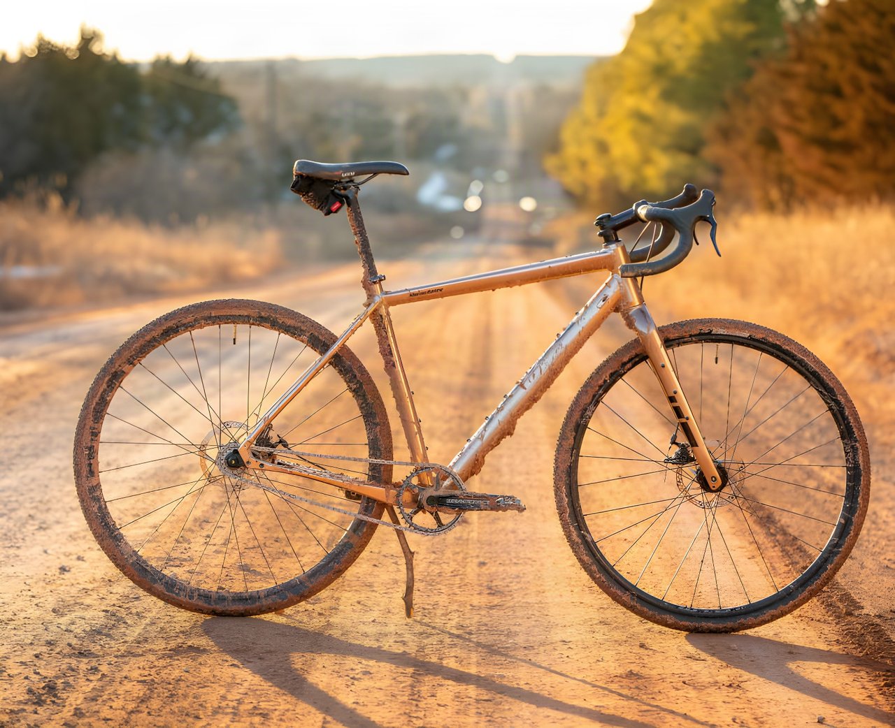 A mud-splattered gold Salsa Stormchaser singlespeed gravel bike stands on an Oklahoma dirt road at sunset.