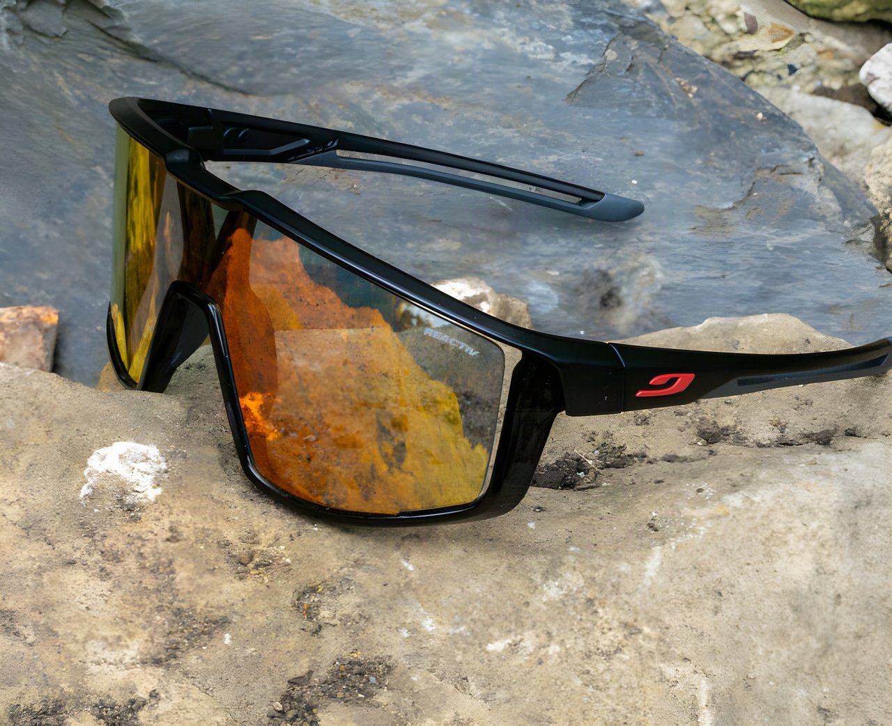 Julbo Fury Reactiv sunglasses with a black frame and orange mirrored photochromic lens resting on rock.