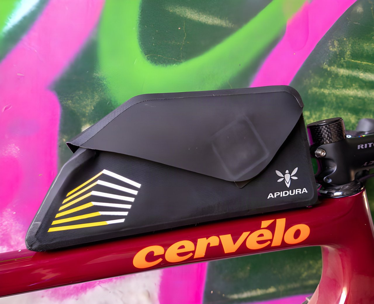 Apidura Racing bolt-on top tube bag with its flip-top cover on a maroon Cervélo frame against a graffiti wall.