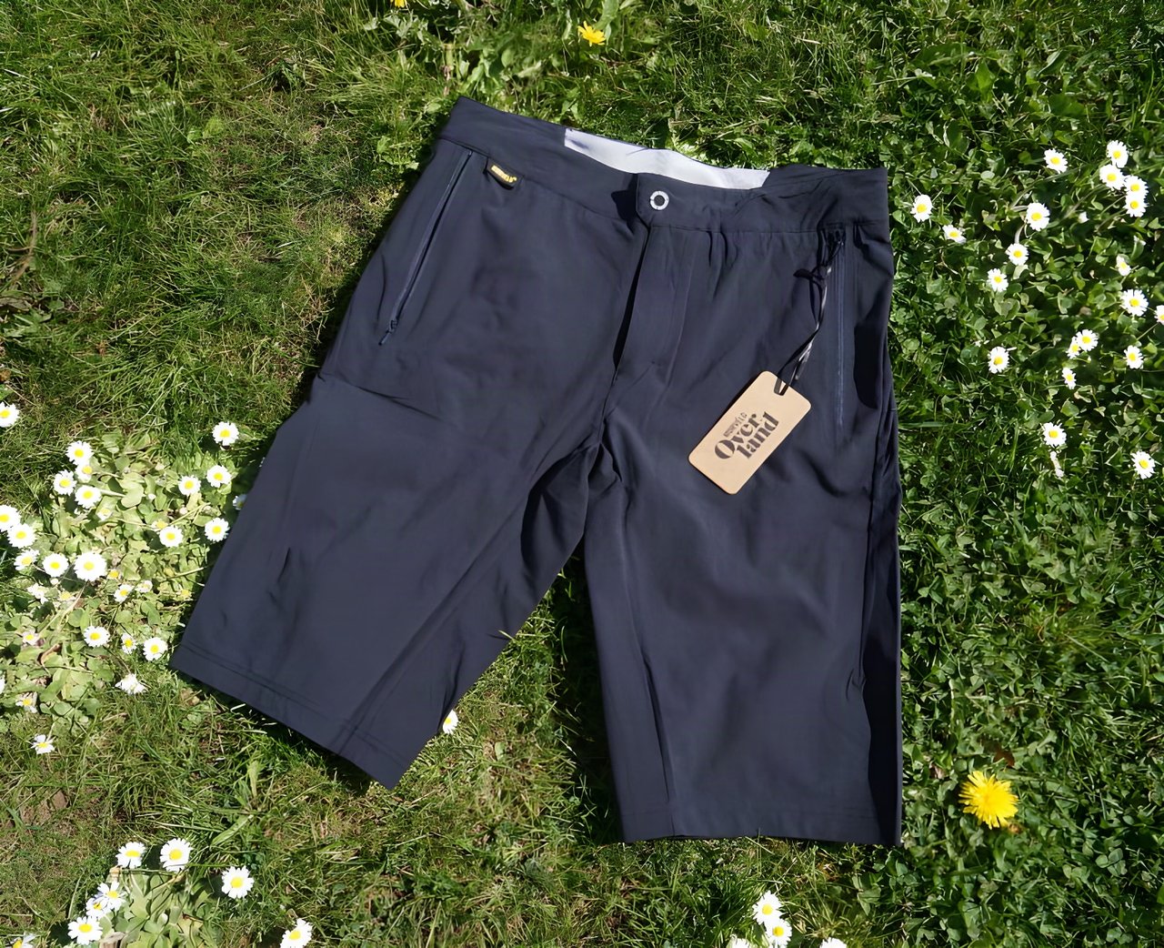 Overland Selector shorts in dark grey laid on grass among daisies, with the cardboard Morvélo Overland tag attached.