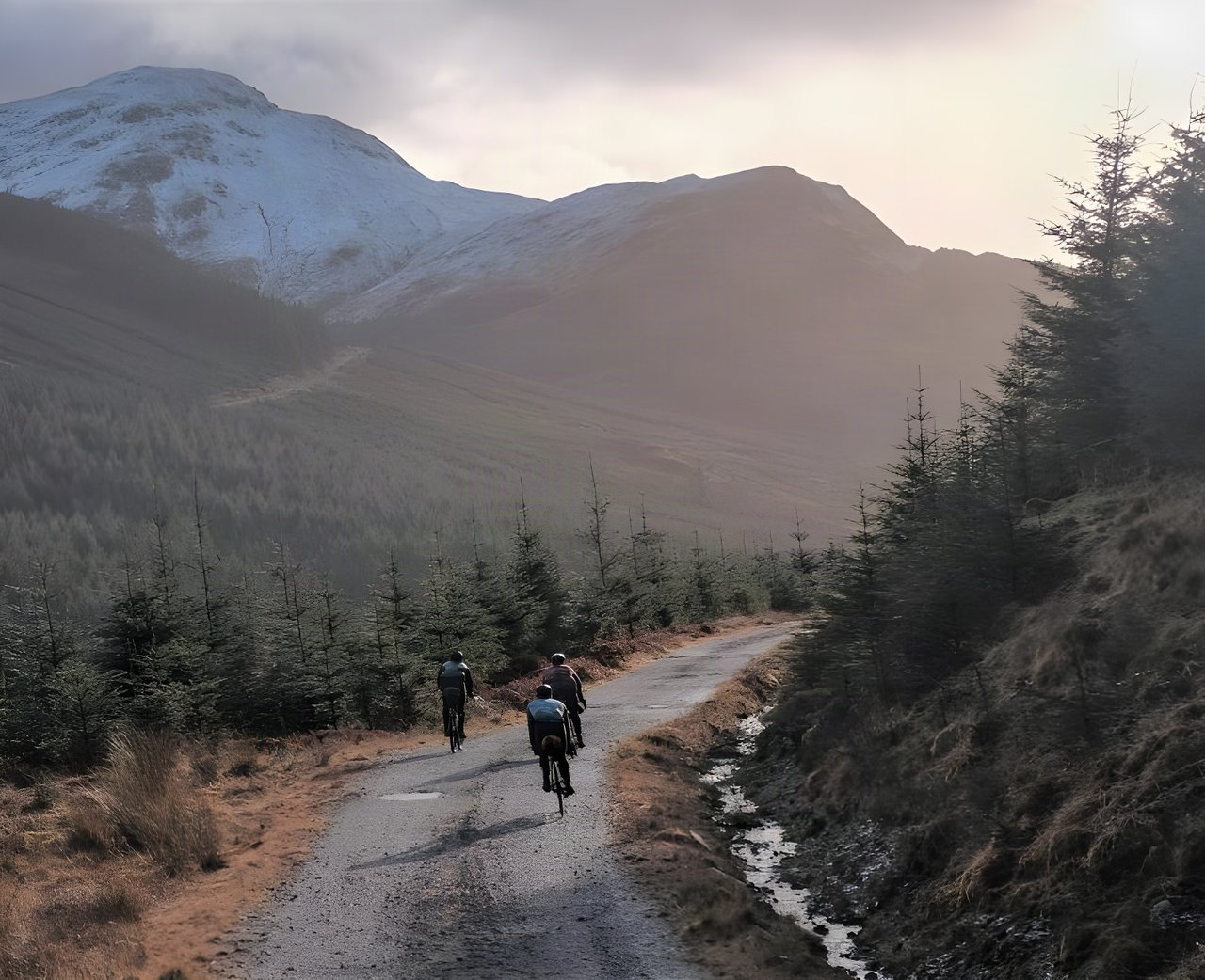 Three riders on a forest road below snow-dusted hills on the bikepacking trip to Mark Cottage bothy in Scotland.