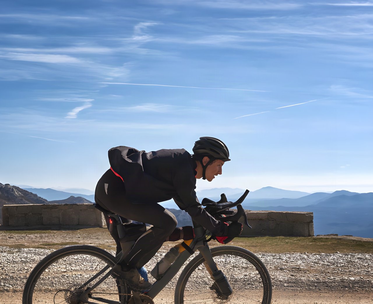 Garmin Edge Plus launch image: a rider on a gravel bike crosses a stony mountain road beside a low wall under blue sky.