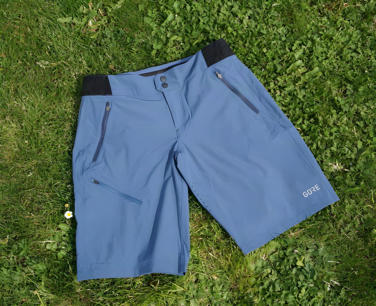 GORE C5 Women's Shorts in deep water blue laid on grass, with zipped hip pockets, poppers and black waistband tabs.