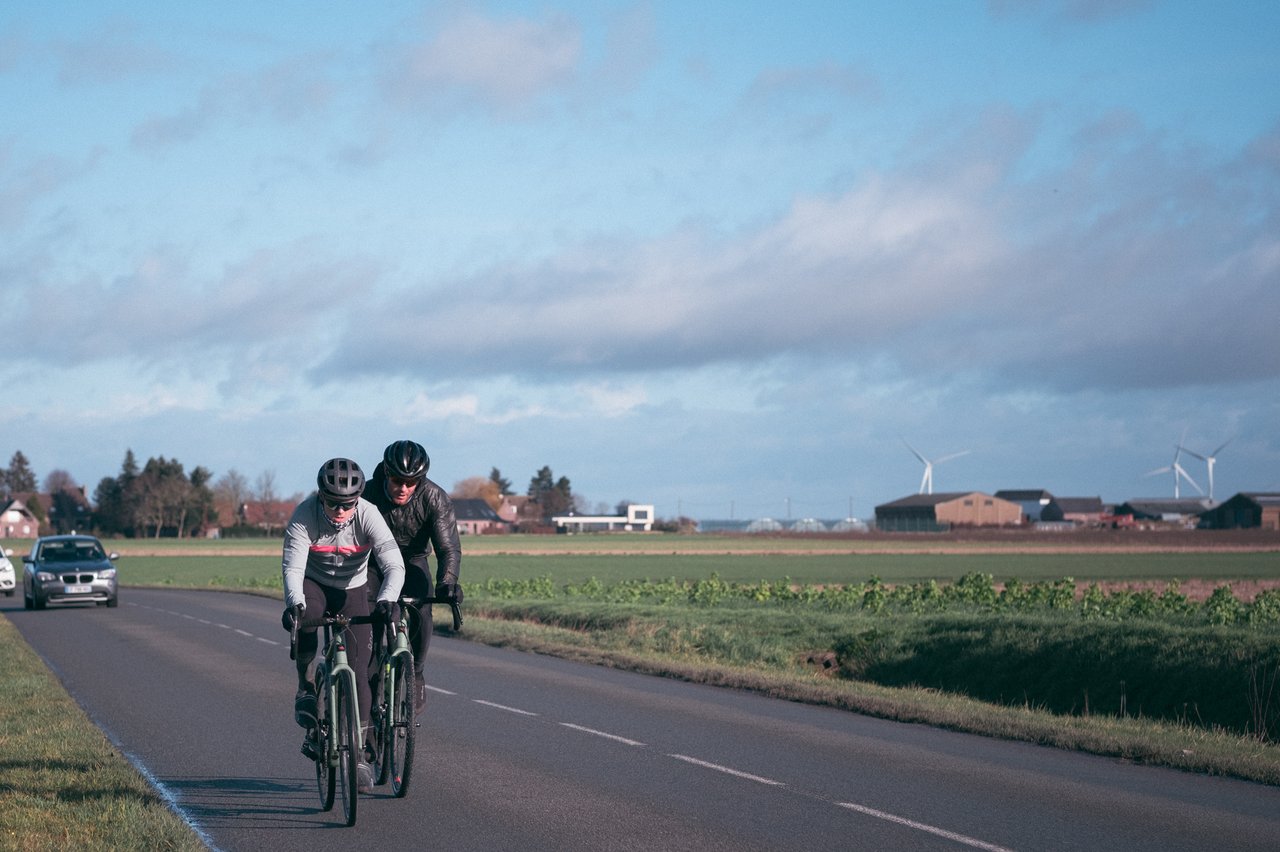 Two riders on Santa Cruz Stigmata road bikes pass flat farmland and wind turbines during the Creepy Fatigue trip in Belgium.