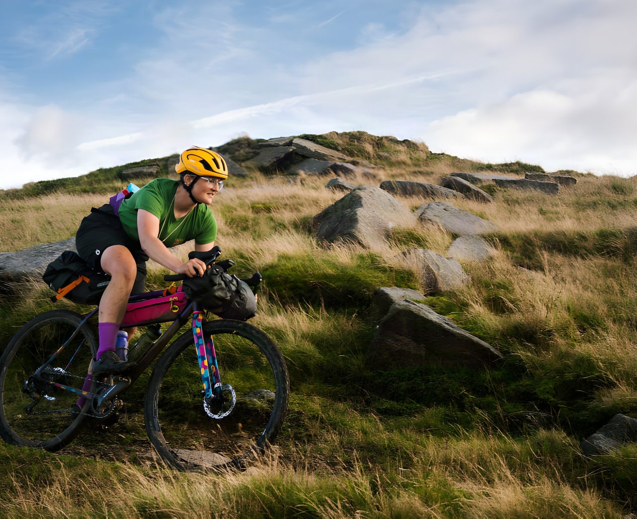 Katherine Moore rides her custom steel Mercredi, loaded with bikepacking bags, along a rocky moorland hillside.