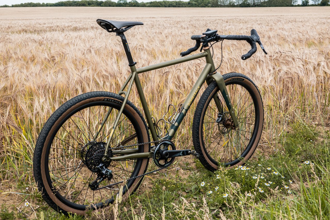Bombtrack Hook EXT gravel bike in matte olive with tan-wall Teravail tyres, parked beside a ripe barley field.