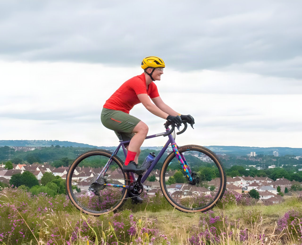 Katherine Moore rides a purple gravel bike through heather in Velocio TRAIL kit, red tee and khaki shorts, above a town.