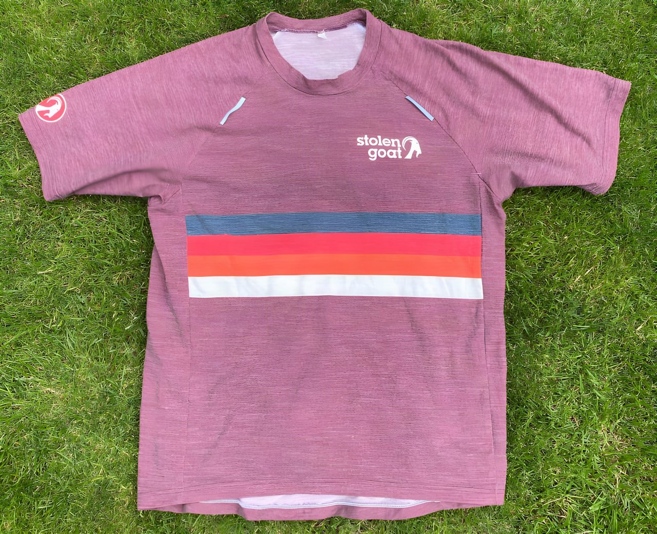 Stolen Goat Merino Gravel Jersey in washed-out red with retro stripes across the chest, laid flat on grass.
