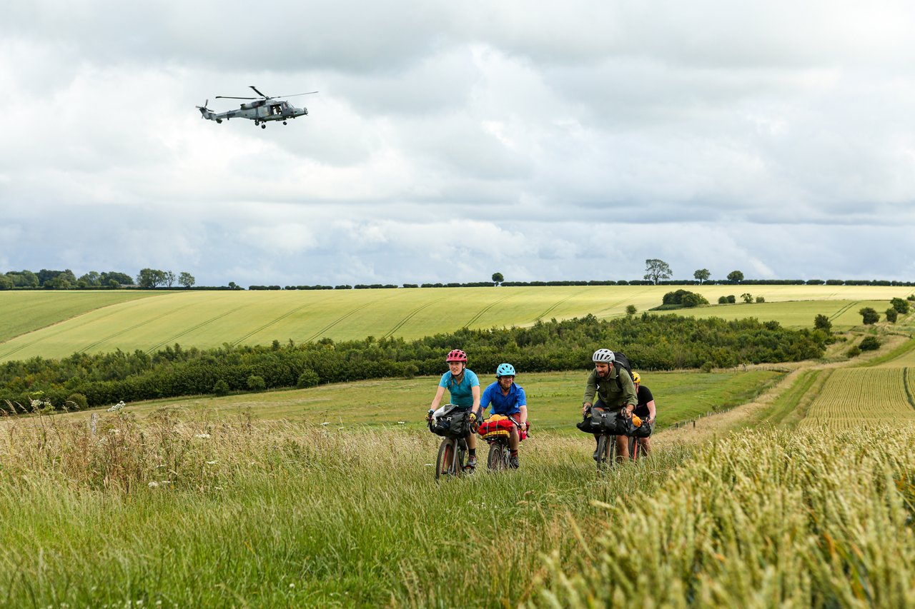 Bikepackers ride a grassy track on the King Alfred's Way as a military helicopter passes over the Wessex farmland.