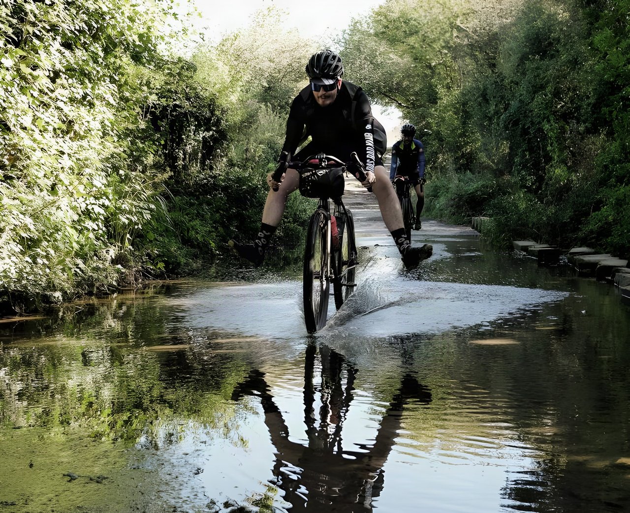 AUTMNL ADVNTR rider splashes through a flooded lane on a bike with a handlebar bag, a second rider following.