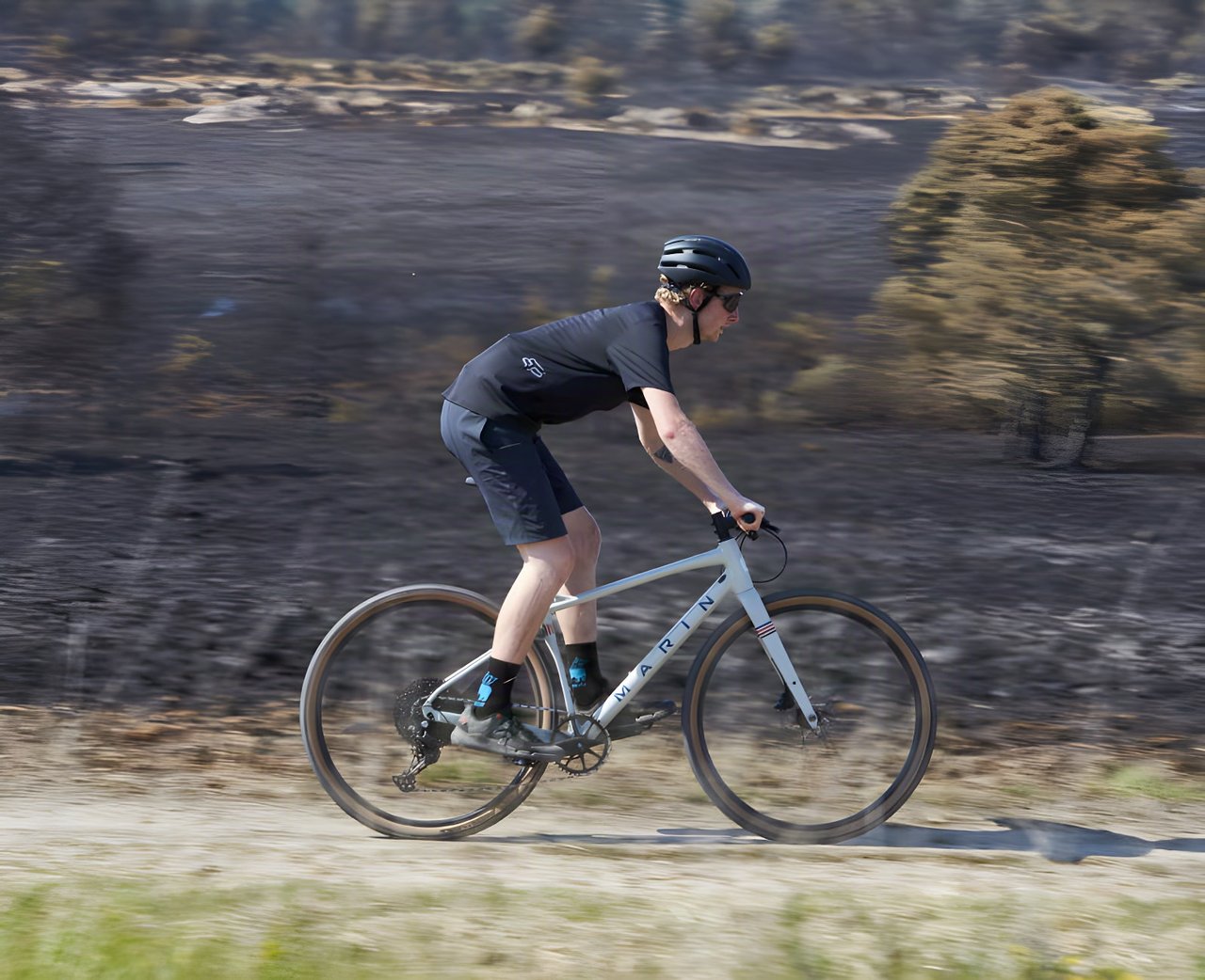 Rider on the off-white flat-bar Marin DSX gravel bike speeds along a dry dirt track past scorched heathland.