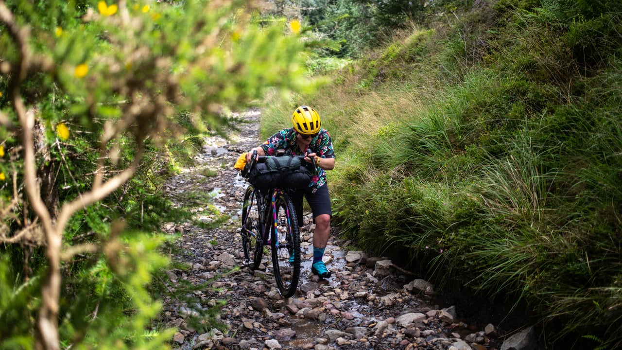 Rider pushes a loaded gravel bike with a handlebar bag up a rocky, stream-worn track on a hike-a-bike section.