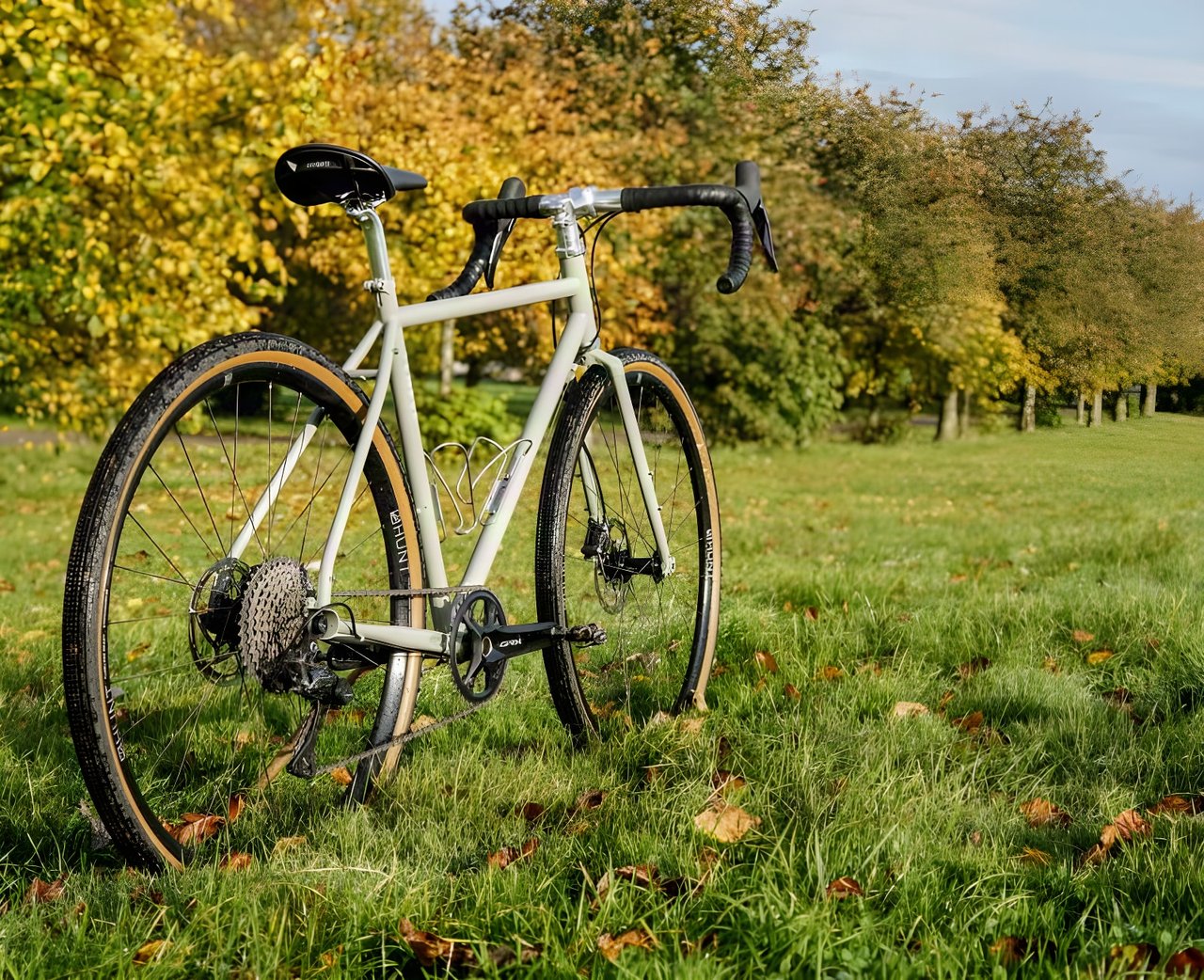 Temple Adventure Disc 1 steel gravel bike in Lichen Green with tan-wall tyres on autumn grass beside trees.