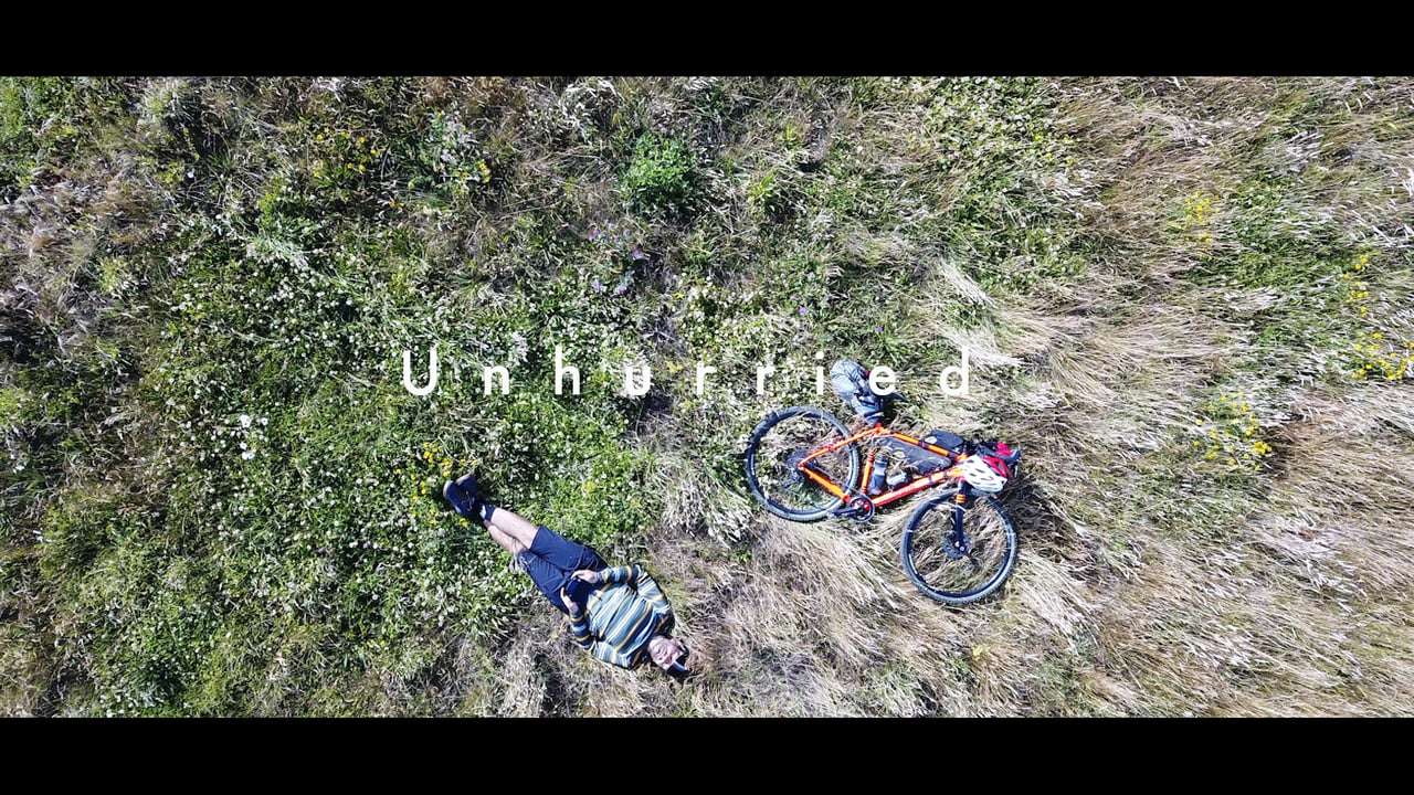 Overhead frame from the film Unhurried: a rider lies in long grass beside a loaded bikepacking bike.