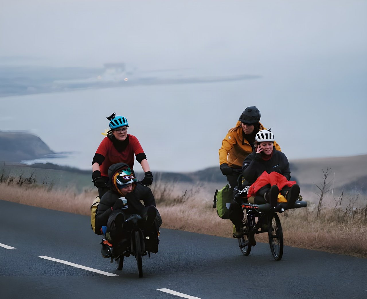 Four Adventure Syndicate riders share two cargo bikes on a coastal road above a grey sea during the Resolution Race.