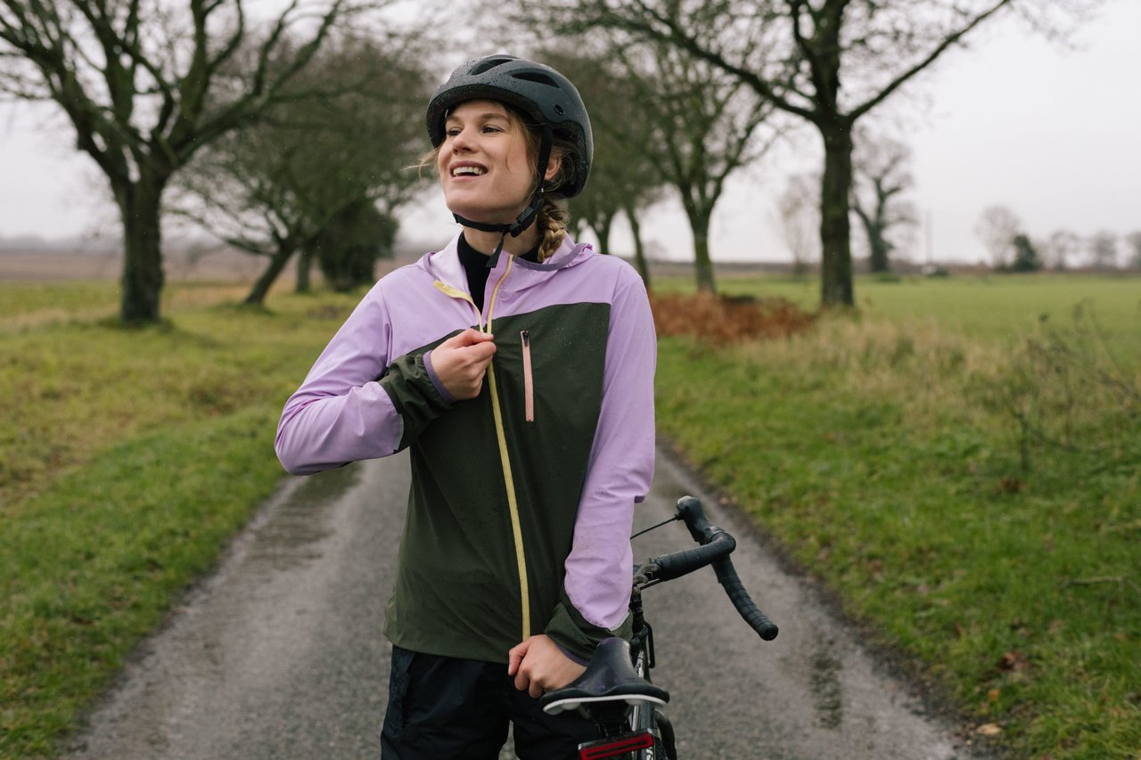 Rider in a lilac and green IRIS Escape Jacket unzips it beside a drop-bar bike on a wet country lane.