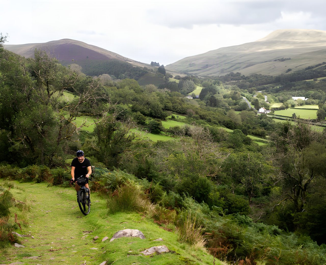 Bikepacker rides a grassy track above a green valley and hills in Wales, on one of the UK's bikepacking routes.