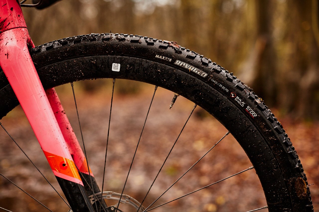 Mud-splashed Maxxis Ravager SilkShield gravel tyre on a pink gravel bike's front wheel in autumn woodland.