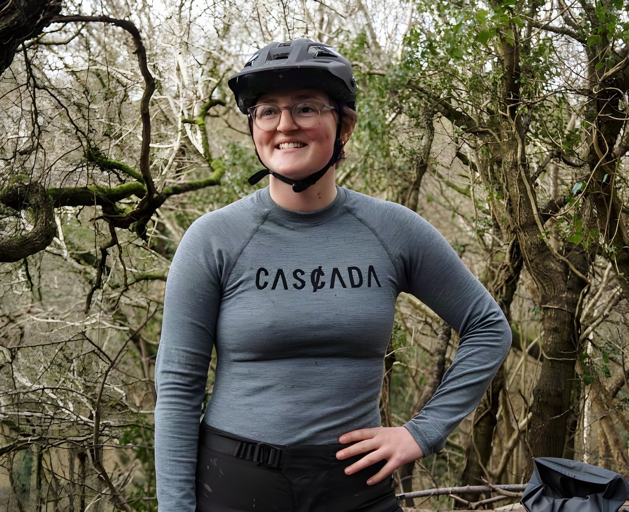 Rider in a grey long sleeve Cascada merino baselayer and helmet, hand on hip in winter woodland.