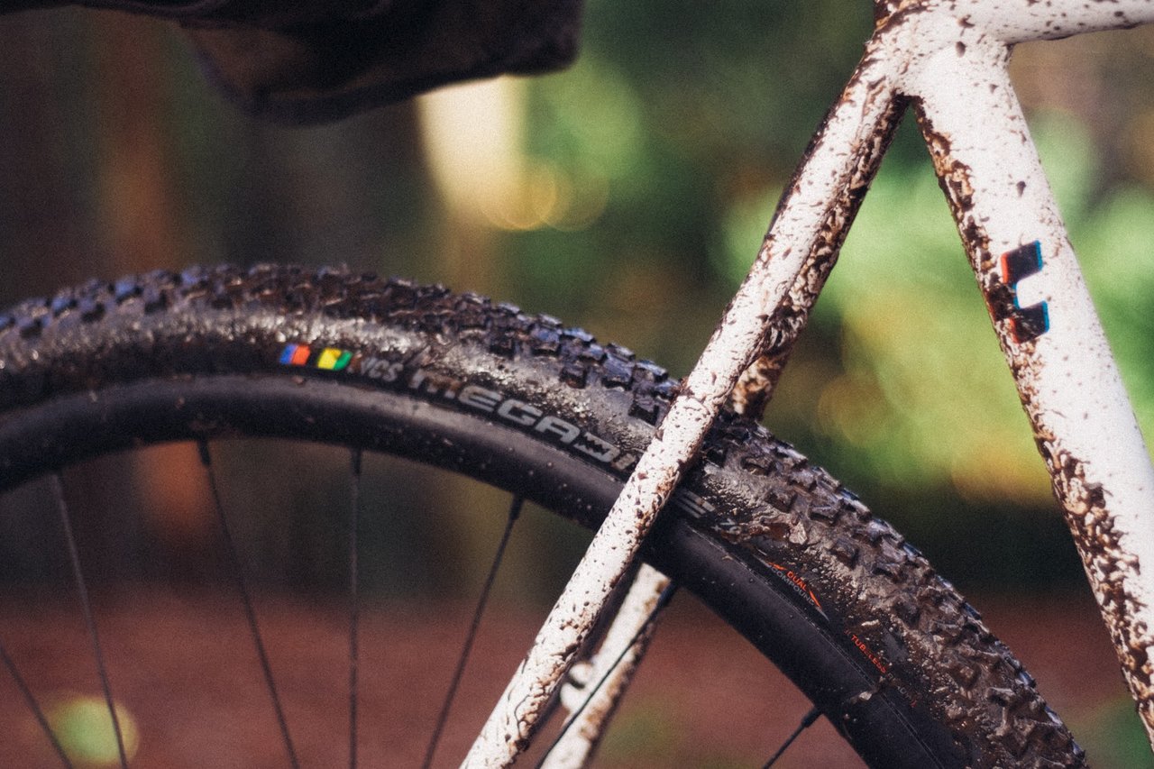 Mud-splattered Ritchey MegaBite WCS cyclo-cross tyre beside a white frame tube, close up in woodland.