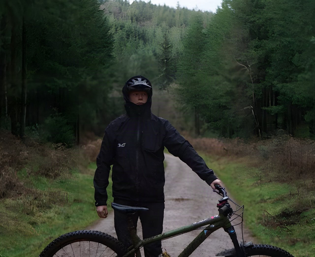 Rider in a black TSG Drop Rain Jacket and helmet stands with a mountain bike on a forest track.