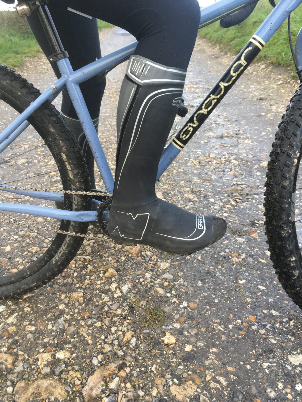 SPATZ GravlR overshoe covering a rider's lower leg, foot on the pedal of a blue bike on a wet gravel track.