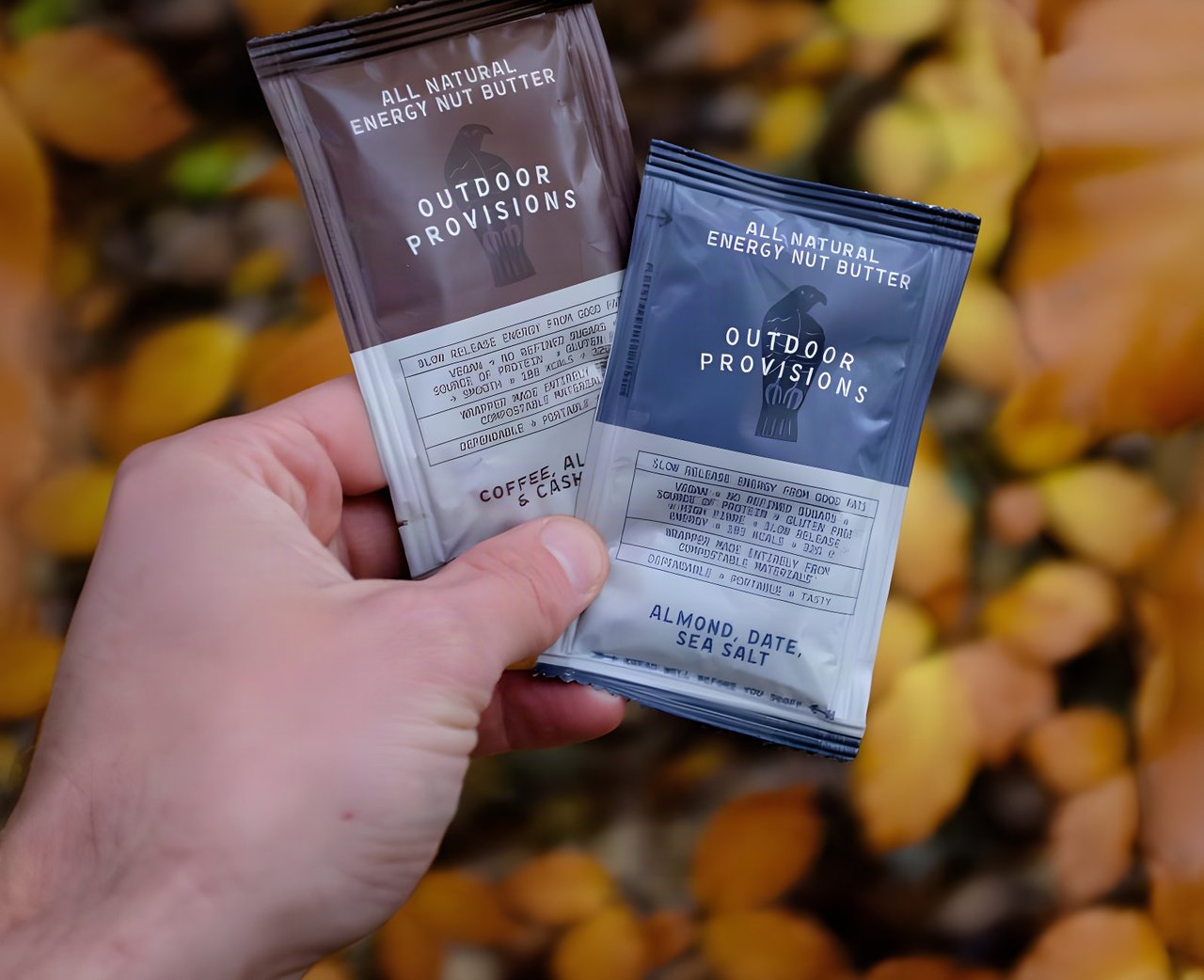 Hand holding two Outdoor Provisions energy nut butter sachets against autumn leaves.
