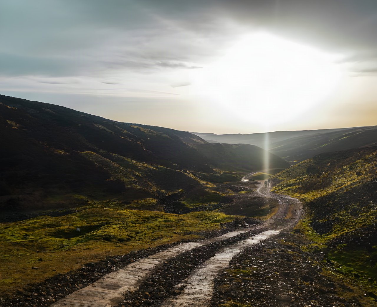 Gravel track winding through a moorland valley towards a low sun, with two distant riders, from Restrap's route catalogue.
